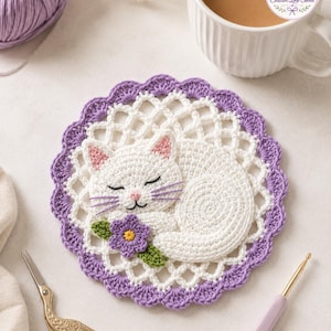 May include: A round crochet coaster featuring a white sleeping cat with pink inner ears and purple whiskers. The cat rests on a white lattice-patterned base with a scalloped purple border. A small purple flower with a yellow center and green leaves is attached.