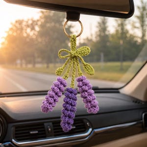 May include: A crochet lavender flower car accessory hanging from a rearview mirror. The item features three purple textured flower stems attached to a green stem with a small bow and two leaves, all connected to a gold-toned metal ring.