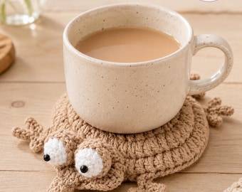 May include: A light brown crocheted coaster shaped like a frog with large white eyes and black pupils, supporting a speckled cream-colored ceramic mug filled with light brown liquid. The logo Charm Loop Crochet is visible in the upper right corner.