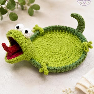 May include: A round, green crocheted tray shaped like a frog with a wide-open mouth, a red tongue, and two white eyes with black pupils. The frog has four small limbs and a curved tail extending from the circular base, crafted with yarn.
