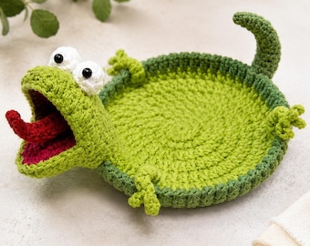 May include: A round, green crocheted tray shaped like a frog with a wide-open mouth, a red tongue, and two white eyes with black pupils. The frog has four small limbs and a curved tail extending from the circular base, crafted with yarn.