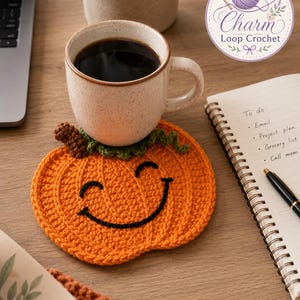 May include: A bright orange crochet pumpkin coaster with a black stitched smiling face, a green stem, and a brown textured top. The coaster sits under a speckled ceramic mug filled with dark coffee, next to a spiral notebook and a black pen.