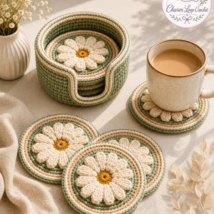 Puede incluir: Un juego de posavasos redondos de ganchillo con un diseño de margarita blanca y centro amarillo, sobre un fondo verde salvia y crema. Los posavasos se guardan en un soporte de ganchillo a juego, con uno de ellos sosteniendo una taza de cerámica.