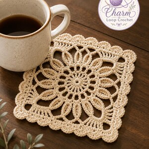 May include: A square, cream-coloured crochet coaster featuring a central floral motif with intricate openwork loops and a scalloped edge. A ceramic mug filled with dark liquid sits partially on the coaster, which displays a circular logo reading Charm Loop Crochet.