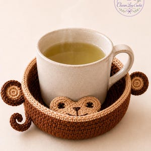 May include: A beige ceramic mug filled with green liquid sits in a handmade crochet monkey coaster. The coaster is brown and tan, with a monkey face and ears. The Charm Loop Crochet logo is in the upper right corner.