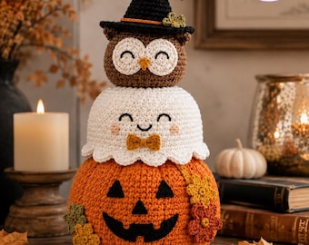 May include: A stacked crochet decoration featuring an orange pumpkin base with a black jack-o'-lantern face, a white ghost middle section with a small orange bow tie, and a brown owl top wearing a black witch hat with a small green flower.