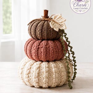 May include: A stacked set of three crocheted pumpkins in brown, dusty rose, and cream. The top pumpkin features a brown stem, a cream crocheted leaf, and a long, curly green vine. The pumpkins have a ribbed texture and are arranged vertically.