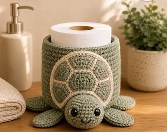 May include: A sage green crocheted turtle toilet paper holder with a white shell pattern and black plastic eyes. The cylindrical body holds a standard roll of white toilet paper. The turtle has four flat flippers and a small head extending forward.