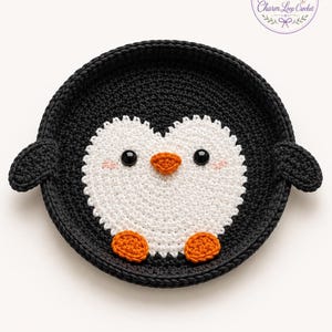 Puede incluir: Un posavasos de pingüino de ganchillo hecho a mano. El posavasos presenta un borde negro y una cara de pingüino blanca con ojos negros, un pico naranja y pies naranjas. El diseño incluye el texto "Charm Loop Crochet".