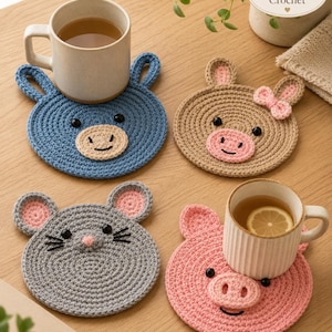Puede incluir: Cuatro posavasos redondos de ganchillo con caras de animales: un oso azul, un conejo beige con un lazo rosa, un ratón gris y un cerdo rosa. Cada posavasos tiene ojos y detalles faciales negros bordados. Dos tazas de cerámica están encima.