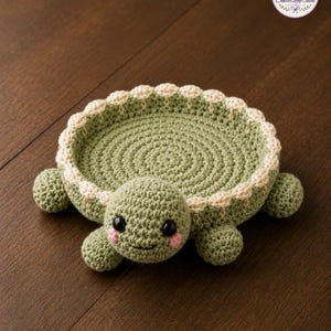 May include: A round, sage green crochet turtle tray with a cream-colored scalloped border. The turtle features a flat, circular shell, four small legs, and a head with black plastic eyes and pink stitched cheeks. The design is textured and handmade.