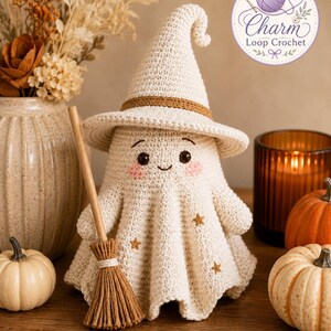 May include: A cream-colored crocheted ghost figurine wearing a pointed hat with a brown band, featuring embroidered pink cheeks, black eyes, and small gold stars on its body. The ghost holds a small wooden broomstick with brown bristles.