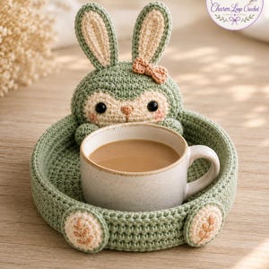May include: A handmade crochet bunny tray in shades of green and beige, holding a white ceramic mug filled with coffee. The tray features a bunny face and ears, with a small bow. The Charm Loop Crochet logo is visible in the upper right corner.