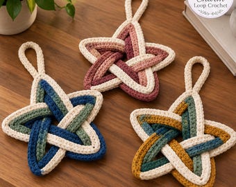 May include: Three star-shaped crochet ornaments with hanging loops. One is pink and cream, one is blue and green with cream, and one is mustard yellow and green with cream. Each features a woven, layered design with a textured yarn finish.