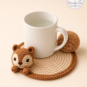 Puede incluir: Un posavasos de ganchillo hecho a mano con un diseño de ardilla marrón y una cola de pompón. El posavasos es de color beige y marrón, con una taza blanca encima. El logotipo "Charm Loop Crochet" está en la esquina superior derecha.