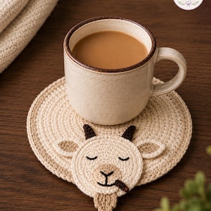 May include: A beige ceramic mug filled with coffee sits on a handmade crochet coaster. The coaster features a goat face design with closed eyes, horns, and a beard. The coaster is beige and tan, with a circular shape.