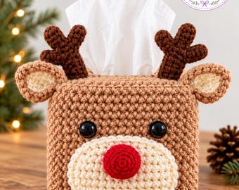 May include: A square tissue box cover crocheted in light brown yarn, featuring a reindeer face with dark brown antlers, cream-colored ears, black plastic eyes, and a bright red nose. A white tissue is pulled through the top opening.