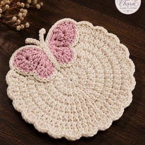May include: A round, cream-colored crochet coaster with a scalloped edge and a decorative butterfly motif attached to the top. The butterfly features dusty pink wings outlined in cream yarn. A circular logo in the corner reads Charm Loop Crochet.