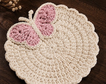 May include: A round, cream-colored crochet coaster with a scalloped edge and a decorative butterfly motif attached to the top. The butterfly features dusty pink wings outlined in cream yarn. A circular logo in the corner reads Charm Loop Crochet.