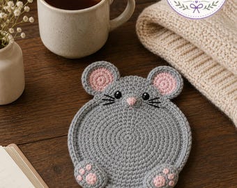 May include: A handmade gray crochet mouse coaster with pink ears and paws. The coaster is round and sits on a wooden surface next to a mug of tea, a small vase of flowers, and a knitted item. The Charm Loop Crochet logo is visible.