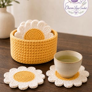 May include: A set of crochet daisy coasters with white petals and a yellow center, stored in a matching yellow crochet basket. One coaster holds a beige ceramic cup filled with tea. The logo Charm Loop Crochet is visible in the top right corner.