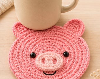 May include: A pink crocheted pig coaster with black button eyes, a pink snout, and a smiling mouth. A beige mug with a brown liquid sits on the coaster. The coaster has two small ears and is on a light wooden surface.