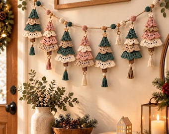 May include: A decorative garland featuring seven crocheted Christmas trees in shades of dusty pink, cream, and dark teal. The trees are strung on a cord with wooden beads, small stars, and hanging tassels in matching colors.