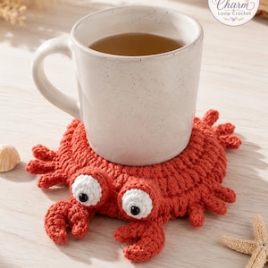 May include: A round, orange, crocheted crab coaster with two white, circular eyes featuring black pupils. The crab has small crocheted legs and claws extending from its body. A light-colored ceramic mug filled with tea sits on top of the coaster.