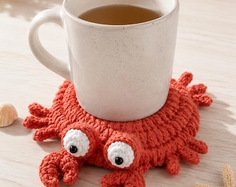 May include: A round, orange, crocheted crab coaster with two white, circular eyes featuring black pupils. The crab has small crocheted legs and claws extending from its body. A light-colored ceramic mug filled with tea sits on top of the coaster.