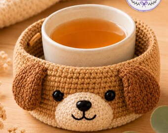 May include: A handmade crochet dog-shaped cup cozy in shades of brown and cream, with black button eyes and a black nose. A white ceramic cup filled with tea sits inside the cozy. The cozy is designed to keep beverages warm.