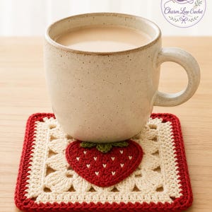Könnte beinhalten: Ein quadratischer Häkeluntersetzer mit einem cremefarbenen Blumenmuster, einer zentralen roten Erdbeerapplikation und einem roten Rand. Eine gesprenkelte beige Keramiktasse mit einem hellbraunen Getränk steht auf dem Untersetzer.