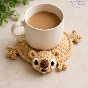 May include: A beige crocheted coaster shaped like a koala with a round body, small ears, and a dark brown nose, supporting a speckled off-white ceramic mug filled with light brown coffee. The logo Charm Loop Crochet is visible in the top right corner.