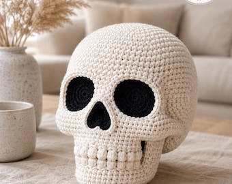 May include: A cream-colored crocheted skull with deep black eye sockets and a black nasal cavity. The skull features a textured surface created by individual yarn stitches, with a row of rounded, bobble-like stitches forming the teeth.