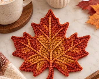 May include: A crochet maple leaf coaster featuring a gradient of autumn colors, transitioning from deep red at the edges to a warm golden yellow in the center, with raised, textured yarn veins radiating from the stem to each leaf point.