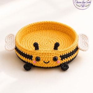 May include: A round, yellow and black crocheted basket shaped like a bee. It features two white crocheted wings on the sides, black antennae, two black plastic eyes, a stitched smile, and small pink cheeks. The interior is yellow.