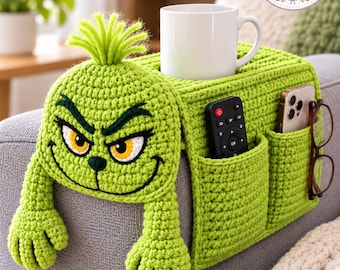 May include: A lime green crochet sofa armrest organizer featuring a character face with yellow eyes, a tuft of hair, and two arms hanging down. It includes two pockets holding a black remote and a smartphone, plus a white mug on top.
