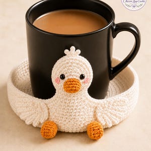 Peut inclure: Une tasse noire remplie d'une boisson brun clair repose sur un dessous de verre en crochet fait à la main, en forme de canard blanc cassé. Le canard a un bec et des pieds orange, avec des joues roses. Le dessous de verre a un design circulaire.
