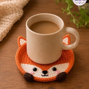 May include: A beige ceramic mug filled with coffee sits on a handmade orange and white crochet fox coaster. The coaster has black eyes, pink cheeks, and brown paws. The mug and coaster are on a wooden surface.
