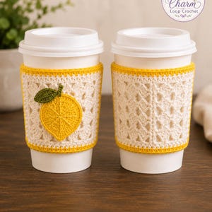 May include: Two white disposable coffee cups with crochet sleeves. One sleeve features a yellow lemon appliqué with a green leaf on a cream-colored square pattern. The other sleeve has a cream-colored square pattern with yellow trim at the top and bottom.