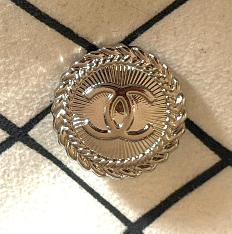 May include: A round, metallic silver-toned button featuring a central embossed interlocking CC logo. The button has a sunburst pattern radiating from the center and a decorative braided rope-style border around the outer edge of the circular design.