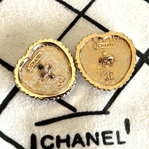 Pode incluir: Dois bot&otilde;es dourados em forma de cora&ccedil;&atilde;o com um design texturizado e a marca "CHANEL" vis&iacute;vel. Os bot&otilde;es t&ecirc;m uma borda ondulada e um poste central para fixa&ccedil;&atilde;o. O fundo &eacute; um tecido branco com texto preto.