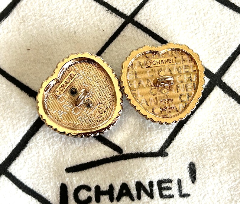 Pode incluir: Dois bot&otilde;es dourados em forma de cora&ccedil;&atilde;o com um design texturizado e a marca "CHANEL" vis&iacute;vel. Os bot&otilde;es t&ecirc;m uma borda ondulada e um poste central para fixa&ccedil;&atilde;o. O fundo &eacute; um tecido branco com texto preto.
