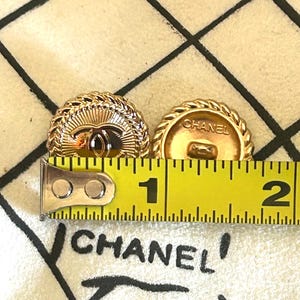 May include: Two round gold-tone buttons featuring a central Chanel logo and a rope-style border. A yellow measuring tape is positioned across the buttons, showing a diameter of approximately 1 inch for each button. The word CHANEL is visible.