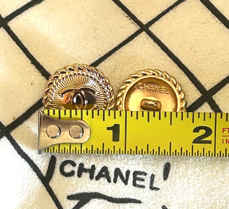 May include: Two round gold-tone buttons featuring a central Chanel logo and a rope-style border. A yellow measuring tape is positioned across the buttons, showing a diameter of approximately 1 inch for each button. The word CHANEL is visible.