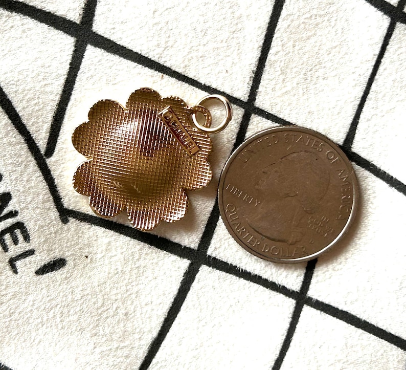 May include: Gold-toned flower-shaped pendant with a textured surface and a small tag that reads "CHANEL". The pendant has a loop for attaching to a chain. A US quarter is next to the pendant.