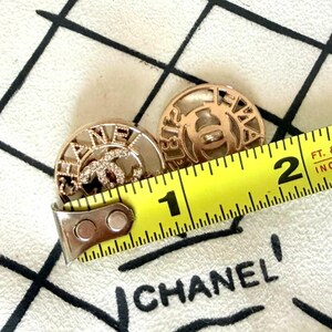 May include: Two round gold-tone buttons featuring the Chanel logo and lettering, with one button accented by small clear crystals. A yellow measuring tape is placed across the buttons, showing a diameter of approximately 1 inch for each button.