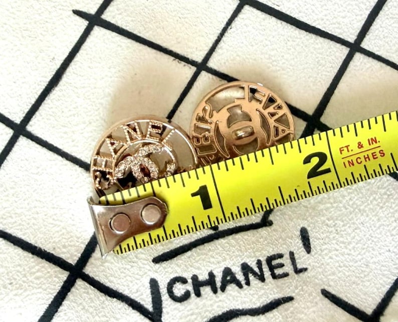 May include: Two round gold-tone buttons featuring the Chanel logo and lettering, with one button accented by small clear crystals. A yellow measuring tape is placed across the buttons, showing a diameter of approximately 1 inch for each button.