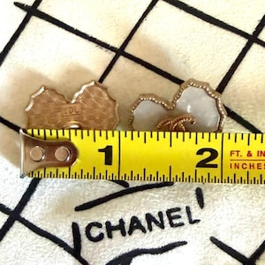 May include: Two heart-shaped buttons with a white center and gold-toned scalloped edges. One button shows a gold interlocking CC logo on the front, while the other shows a textured back with the letters TDL. A yellow measuring tape shows a width of 1.5 inches.