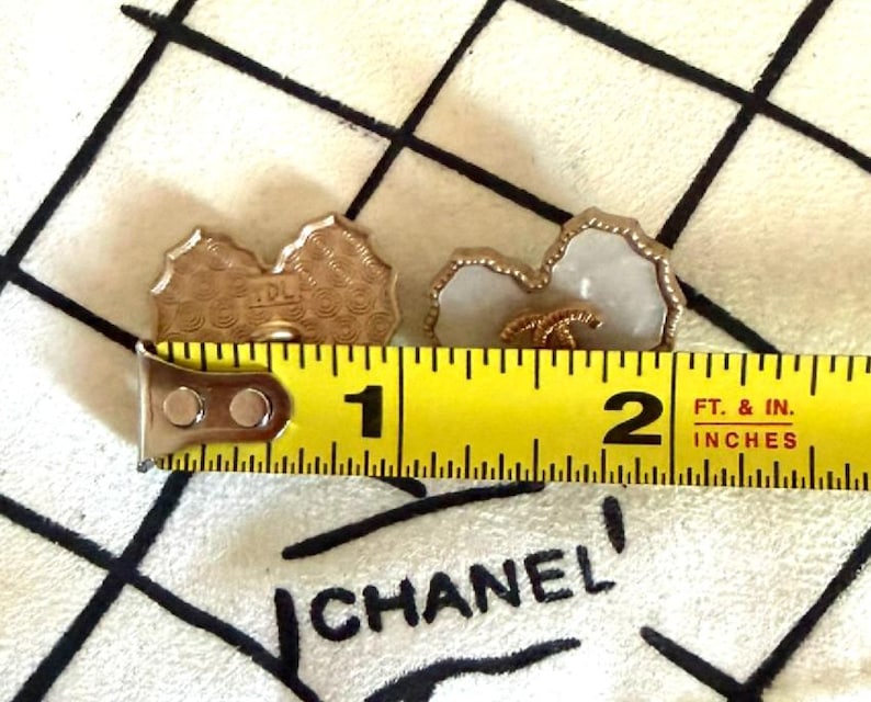 May include: Two heart-shaped buttons with a white center and gold-toned scalloped edges. One button shows a gold interlocking CC logo on the front, while the other shows a textured back with the letters TDL. A yellow measuring tape shows a width of 1.5 inches.