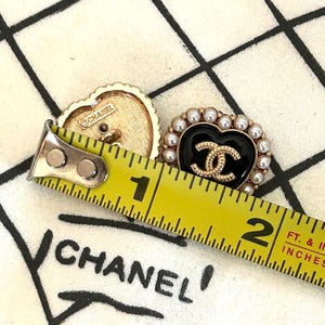 Pode incluir: Dois brincos Chanel em forma de cora&ccedil;&atilde;o. Um &eacute; dourado com o logotipo Chanel e o outro &eacute; preto com um logotipo dourado e rodeado por p&eacute;rolas falsas. Os brincos medem aproximadamente 2,5 cm de comprimento.